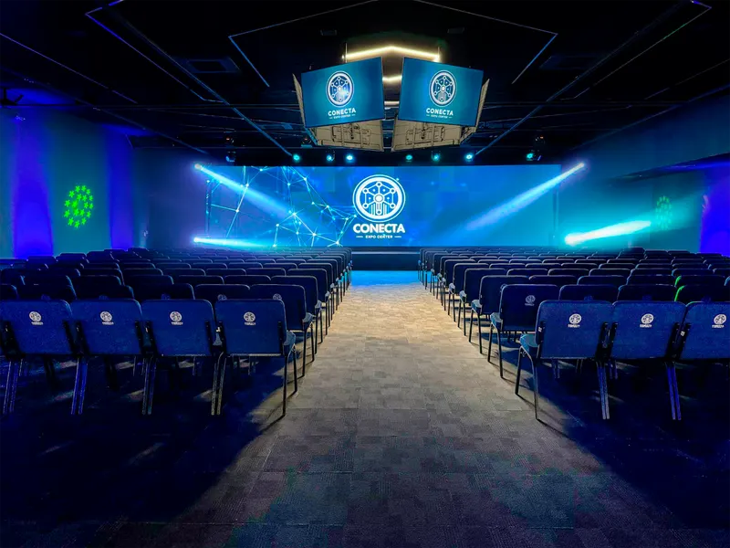 Auditório com fileiras de cadeiras azuis dos dois lados de um corredor central, palco com telão exibindo o logotipo do Conecta Expo Center e duas TVs suspensas, sob iluminação azul e verde.