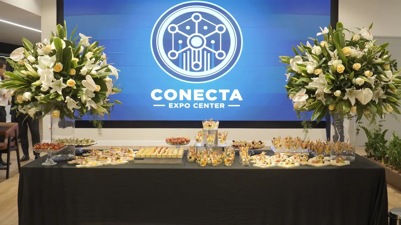 Mesa de coquetel com canapés e arranjos de flores no salão do Coronata, diante do telão com o logo do Conecta