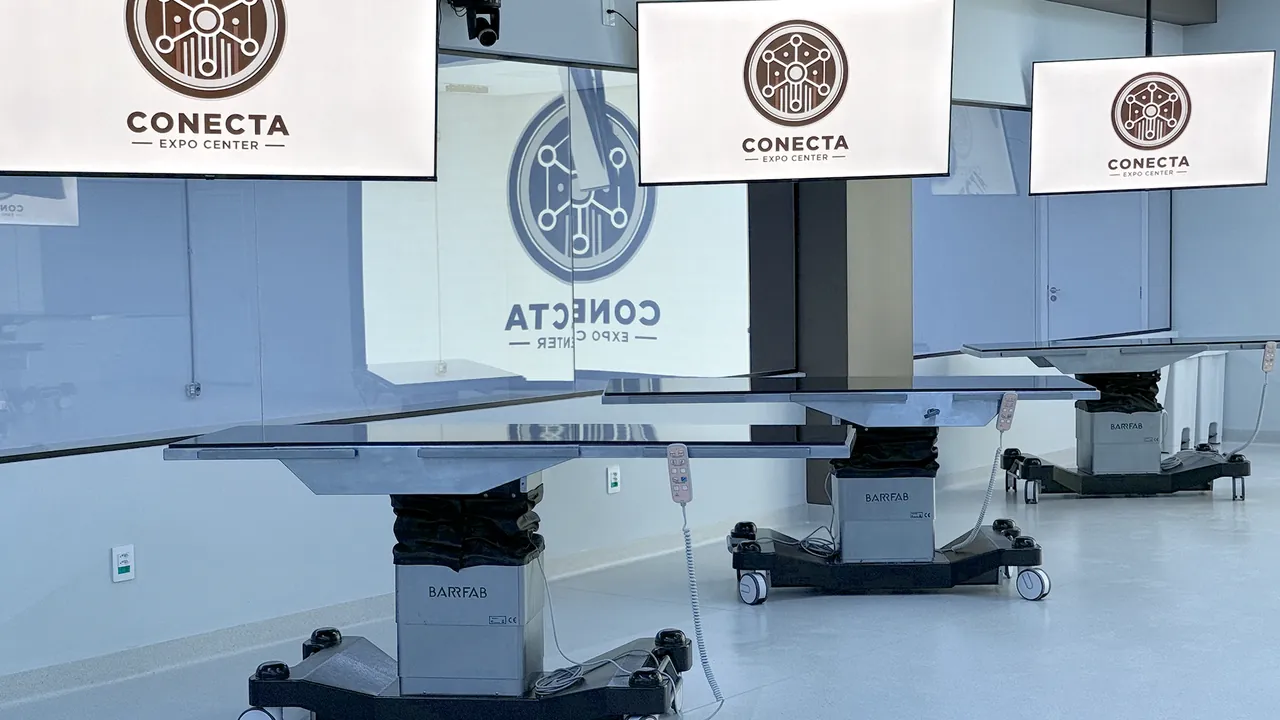 Mesas cirúrgicas do CTC vistas de perto, com o controle preso ao lado e TVs com o logo do Conecta suspensas acima delas