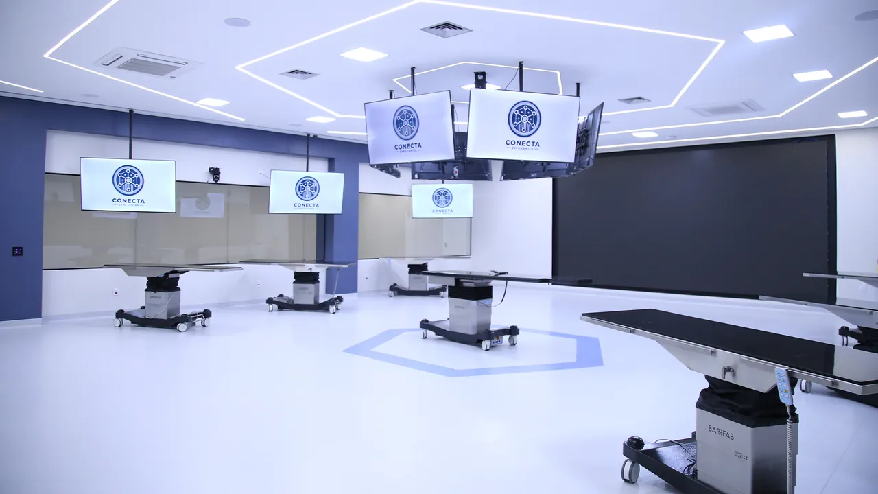 CTC com mesas cirúrgicas sobre bases com rodas, TVs com o logo do Conecta suspensas em trilhos no teto, um painel escuro ao fundo e um hexágono azul desenhado no piso
