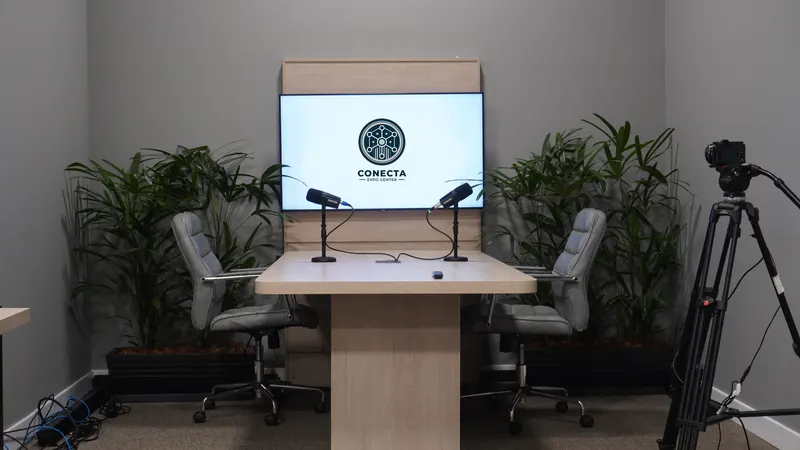 Sala de gravação com mesa de madeira clara, dois microfones em pedestais de mesa e duas cadeiras cinza; ao fundo, TV com o logo do Conecta em painel de madeira, entre vasos de plantas, e uma câmera em tripé à direita