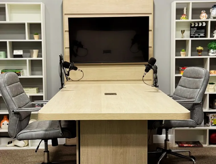Mesa de madeira clara com dois microfones de podcast em braços articulados e duas cadeiras de escritório cinza; atrás, uma TV fixada em painel de madeira e, dos dois lados, estantes brancas com objetos decorativos.