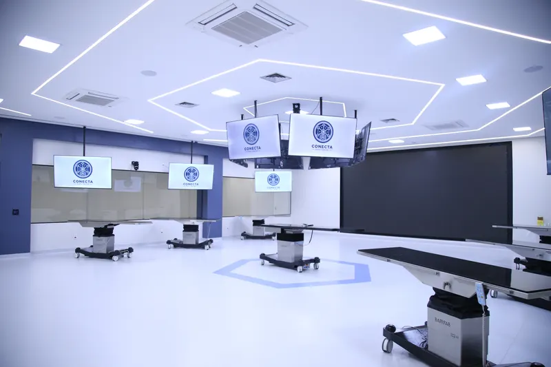 Sala ampla com várias TVs suspensas do teto exibindo o logotipo do Conecta, mesas de tampo escuro sobre colunas com rodas, um painel escuro ao fundo e um hexágono desenhado no piso claro.