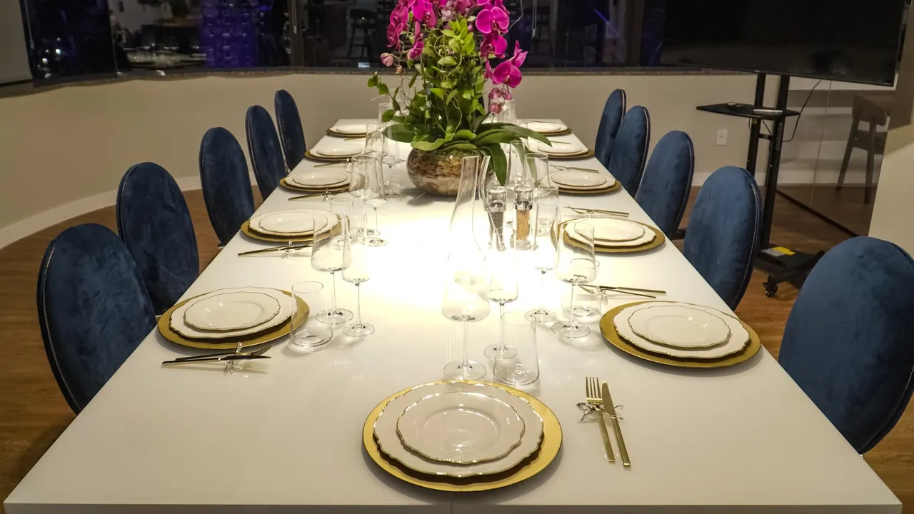 Mesa da Sala Majestic posta com pratos de borda dourada, taças de cristal, orquídeas e cadeiras de veludo azul