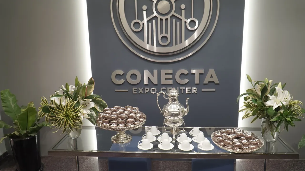 Mesa de café com bule de prata, xícaras, doces e flores diante do logotipo 3D do Conecta Expo Center na recepção