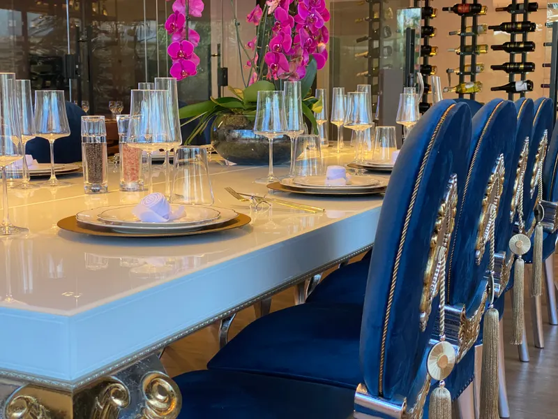 Sala Majestic com mesa posta, pratos, taças e talheres, cadeiras de veludo azul com detalhes dourados, orquídeas rosa e garrafas de vinho ao fundo